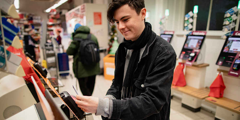 A young man smiling whilst using a self-service checkout machine in a supermarket.