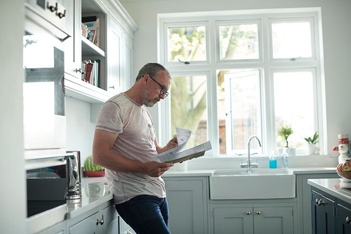 Man stood in a kitchen looking at  a bill that has been received through the post